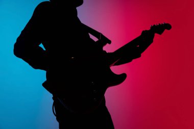 Genç beyaz erkek gitaristin silueti mavi-pembe gradyan stüdyo arka planında neon ışığı altında izole edilmiş.