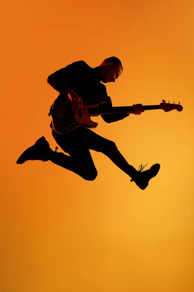 Sylwetka młodego kaukaskiego gitarzysty odizolowanego na pomarańczowym tle studia gradientowego w świetle neonowym — Zdjęcie stockowe