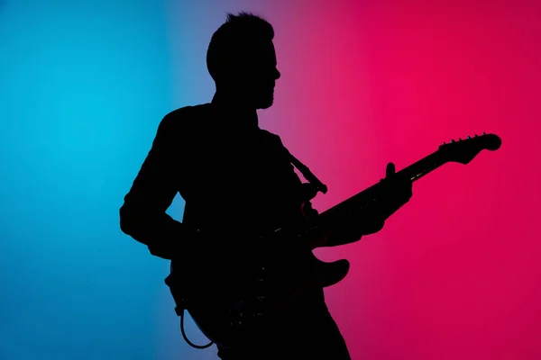 Silhueta de jovem guitarrista masculino caucasiano isolado em fundo estúdio gradiente azul-rosa em luz de néon — Fotografia de Stock