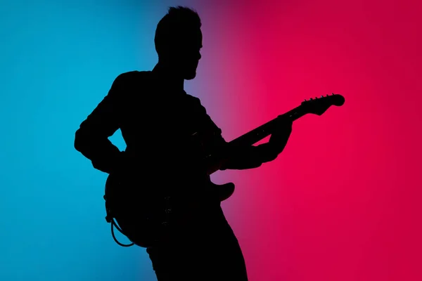 Silhueta de jovem guitarrista masculino caucasiano isolado em fundo estúdio gradiente azul-rosa em luz de néon — Fotografia de Stock