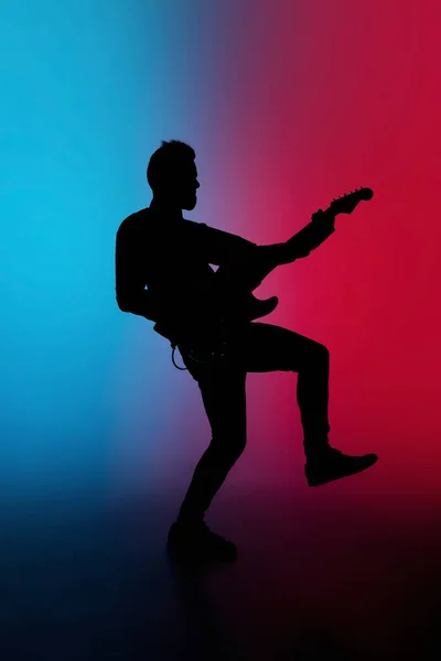 Silhueta de jovem guitarrista masculino caucasiano isolado em fundo estúdio gradiente azul-rosa em luz de néon — Fotografia de Stock
