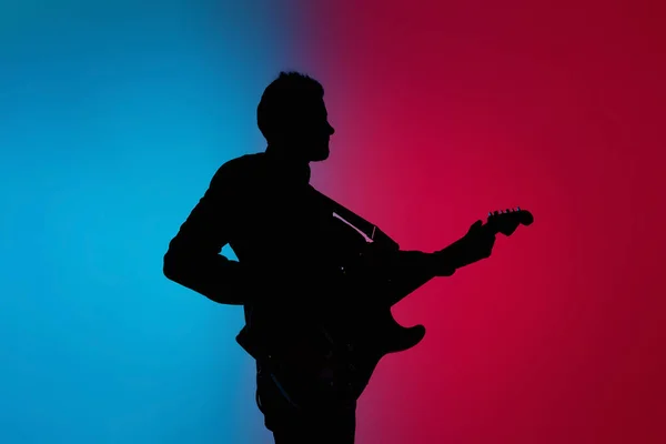 Silhueta de jovem guitarrista masculino caucasiano isolado em fundo estúdio gradiente azul-rosa em luz de néon — Fotografia de Stock