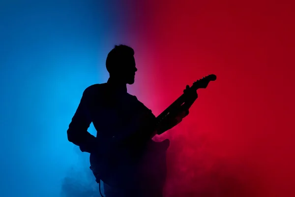 Silhueta de jovem guitarrista masculino caucasiano isolado em fundo estúdio gradiente azul-rosa em luz de néon — Fotografia de Stock