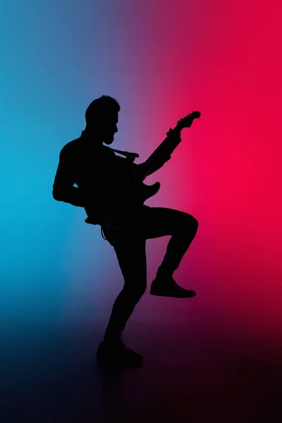 Silhueta de jovem guitarrista masculino caucasiano isolado em fundo estúdio gradiente azul-rosa em luz de néon — Fotografia de Stock