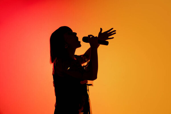 Silhouette of young caucasian female singer isolated on orange gradient studio background in neon light