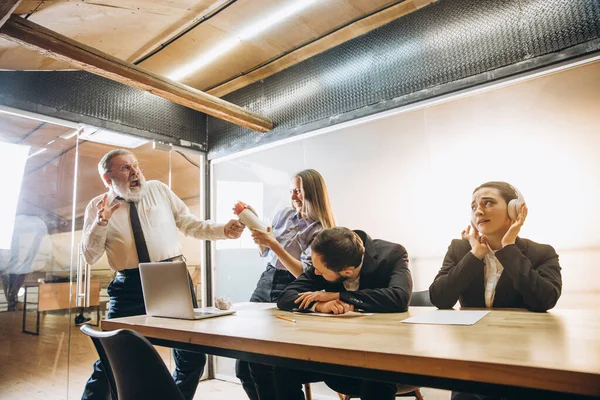 Angry boss with megaphone screaming at employees in office, scared and annoyed colleagues listening at the table