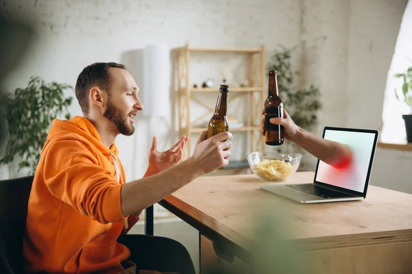 Joven bebiendo cerveza durante la reunión con amigos en videollamada virtual. Distancia reunión en línea, chatear juntos en el ordenador portátil en casa. — Foto de Stock
