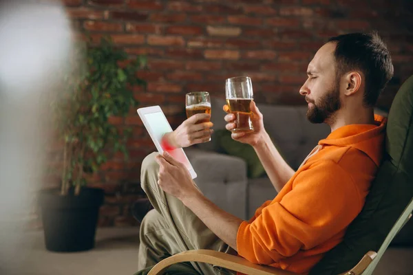 Joven bebiendo cerveza durante la reunión con amigos en videollamada virtual. Distancia reunión en línea, chatear juntos en la tableta en casa. — Foto de Stock