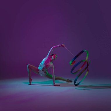 Genç bayan sporcu, ritmik jimnastik sanatçısı mor arka planda, neon ışıklı. Ekipmanlarla pratik yapan güzel bir kız. Gösteride zarafet.