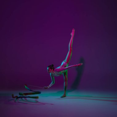 Genç bayan sporcu, ritmik jimnastik sanatçısı mor arka planda, neon ışıklı. Ekipmanlarla pratik yapan güzel bir kız. Gösteride zarafet.