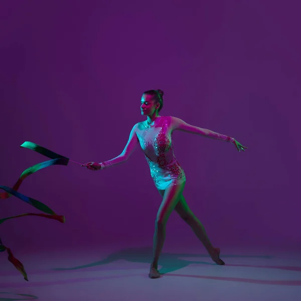 Jovem atleta feminina, artista de ginástica rítmica em fundo roxo com luz de néon. Menina bonita praticando com equipamento. Graça no desempenho. — Fotografia de Stock