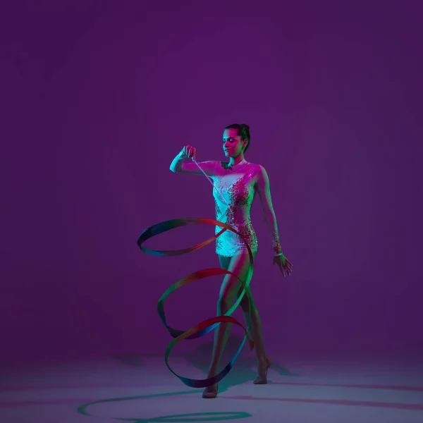 Jonge vrouwelijke atleet, ritmische gymnastiek kunstenaar op paarse achtergrond met neon licht. Mooi meisje dat oefent met apparatuur. Genade in prestaties. — Stockfoto