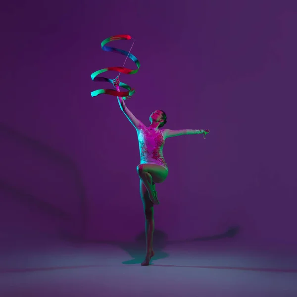 Jovem atleta feminina, artista de ginástica rítmica em fundo roxo com luz de néon. Menina bonita praticando com equipamento. Graça no desempenho. — Fotografia de Stock