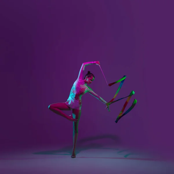 Young female athlete, rhythmic gymnastics artist on purple background with neon light. Beautiful girl practicing with equipment. Grace in performance.