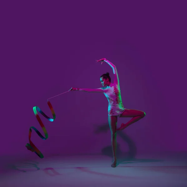 年轻的女运动员,有节奏的体操艺术家,背景为紫色,背景为霓虹灯.漂亮的女孩带着装备练习。优雅的表现. — 图库照片