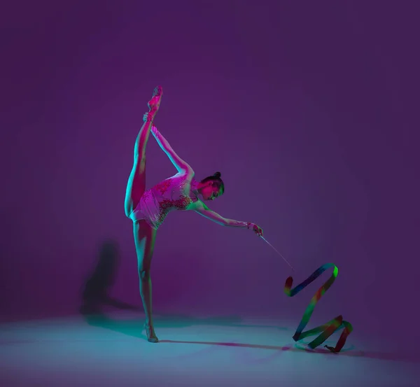 Jonge vrouwelijke atleet, ritmische gymnastiek kunstenaar op paarse achtergrond met neon licht. Mooi meisje dat oefent met apparatuur. Genade in prestaties. — Stockfoto