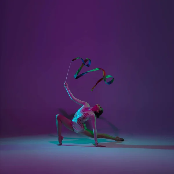 Young female athlete, rhythmic gymnastics artist on purple background with neon light. Beautiful girl practicing with equipment. Grace in performance.