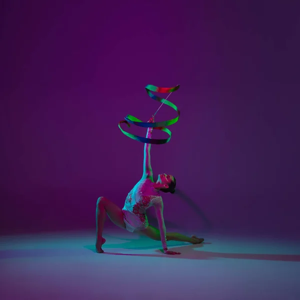 Mladá atletka, rytmická gymnastka na purpurovém pozadí s neonovým světlem. Krásná dívka cvičí s vybavením. Grace v provedení. — Stock fotografie
