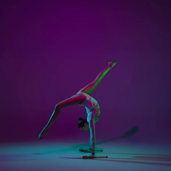 Jonge vrouwelijke atleet, ritmische gymnastiek kunstenaar op paarse achtergrond met neon licht. Mooi meisje dat oefent met apparatuur. Genade in prestaties. — Stockfoto