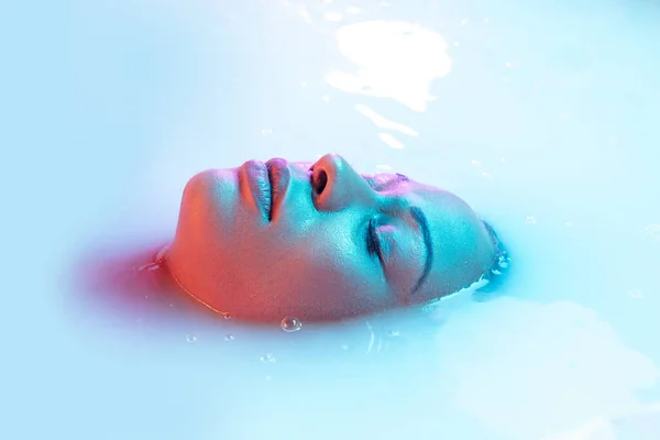 Beautiful female face in the milk bath with soft glowing in blue-pink neon light — Stock Photo, Image