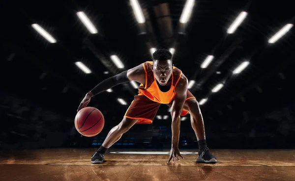 Jugador de baloncesto joven afroamericano en acción y linternas sobre el fondo del gimnasio. Concepto de deporte, movimiento, energía y estilo de vida dinámico y saludable. — Foto de Stock