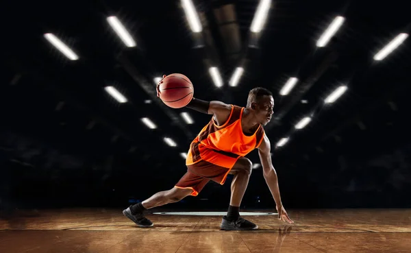 Jugador de baloncesto joven afroamericano en acción y linternas sobre el fondo del gimnasio. Concepto de deporte, movimiento, energía y estilo de vida dinámico y saludable. — Foto de Stock
