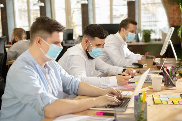 Jovens colegas caucasianos trabalhando juntos em um escritório usando dispositivos modernos e gadgets durante a quarentena. Usando máscaras protetoras — Fotografia de Stock