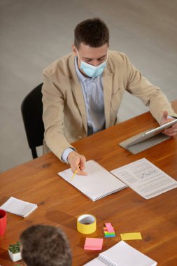 Yaratıcı toplantı sırasında modern cihazlar ve aletler kullanarak bir ofiste karantina sırasında yüz maskeleriyle çalışan yüksek açılı meslektaşlar.