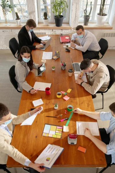 Colleghi con vista ad alto angolo che lavorano insieme in maschere facciali durante la quarantena in un ufficio utilizzando dispositivi e gadget moderni durante riunioni creative — Foto Stock