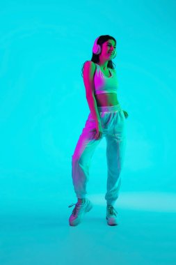Esmer, güzel kadın portresi mavi stüdyo arka planında çok renkli neon ışıkta izole edilmiş. Kulaklıkla dans etmek.