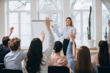 Kadın konuşmacı atölyede sunum yapıyor. Seyirci ya da konferans salonu. Tanınmayan katılımcıların arka görünümü.