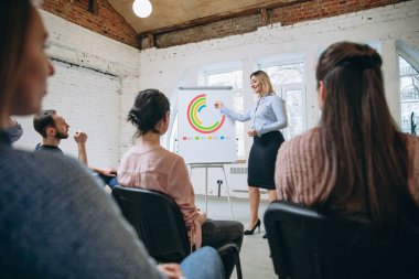 Kadın konuşmacı atölyede sunum yapıyor. Seyirci ya da konferans salonu. Tanınmayan katılımcıların arka görünümü.