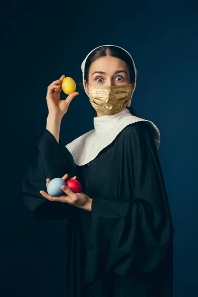 Joven mujer medieval como monja en ropa vintage y máscara dorada sobre fondo azul oscuro. Concepto de comparación de épocas, protección contra el covidio — Foto de Stock