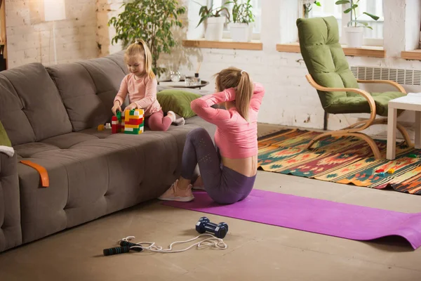 Young woman exercising fitness, aerobic, yoga at home, sporty lifestyle. Getting active while her child playing on the background, home gym.
