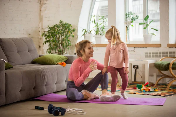Young woman exercising fitness, aerobic, yoga at home, sporty lifestyle. Getting active while child, daughter playing with her, home gym.
