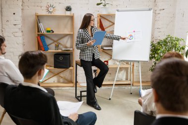 Kadın konuşmacı atölyede sunum yapıyor. Seyirci ya da konferans salonu. Tanınmayan katılımcıların arka görünümü.