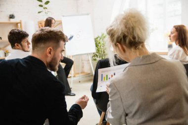 Kadın konuşmacı atölyede sunum yapıyor. Seyirci ya da konferans salonu. Tanınmayan katılımcıların arka görünümü.