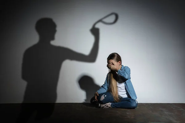 Häusliche Gewalt, Missbrauch. Verängstigtes kleines kaukasisches Mädchen, Opfer sitzt dicht an weißer Wand, auf der der Schatten wütender Väter droht. — Stockfoto