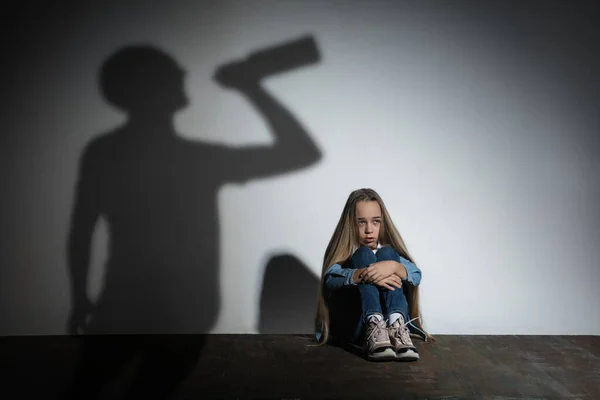 Violencia doméstica, abuso. Pequeña niña caucásica asustada, víctima sentada cerca de la pared blanca con sombra de padre enojado amenazante con adicción al alcohol. — Foto de Stock