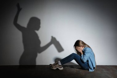 Aile içi şiddet, istismar. Korkmuş küçük beyaz kız, beyaz duvarın yanında oturan, alkol bağımlısı, tehdit eden anne gölgesi olan kurban..