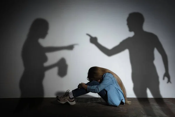 Häusliche Gewalt, Missbrauch. Verängstigtes kleines kaukasisches Mädchen, Opfer, das in der Nähe einer weißen Mauer sitzt, im Schatten wütender Eltern, die mit Alkoholsucht drohen. — Stockfoto