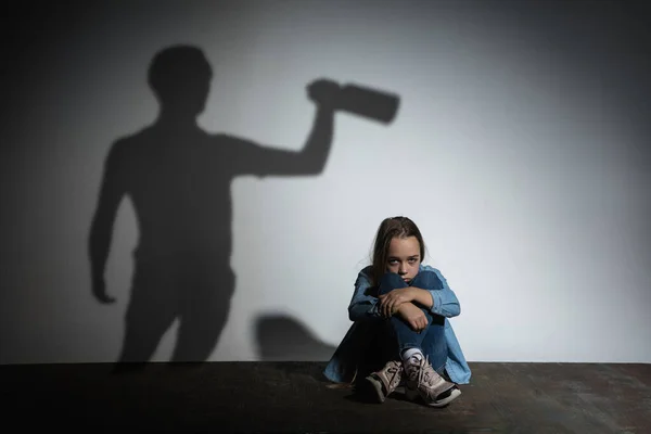 Häusliche Gewalt, Missbrauch. Verängstigtes kleines kaukasisches Mädchen, Opfer sitzt in der Nähe einer weißen Mauer mit dem Schatten eines wütenden Vaters, der mit Alkoholsucht droht. — Stockfoto