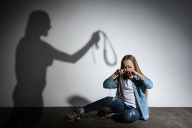 Aile içi şiddet, istismar. Korkmuş küçük beyaz kız, beyaz duvarın yanında oturan kızgın annelerin gölgesiyle tehdit eden kurban..