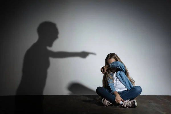 Häusliche Gewalt, Missbrauch. Verängstigtes kleines kaukasisches Mädchen, Opfer sitzt dicht an weißer Wand, auf der der Schatten wütender Väter droht. — Stockfoto