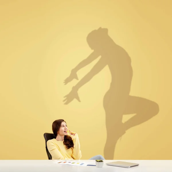 Giovane donna sognando il futuro nel grande sport durante il suo lavoro in ufficio. Diventare una leggenda. Ombra di sogni sul muro dietro di lei. Copyspace. — Foto Stock