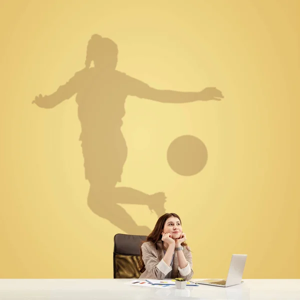 Giovane donna sognando il futuro nel grande sport durante il suo lavoro in ufficio. Diventare una leggenda. Ombra di sogni sul muro dietro di lei. Copyspace. — Foto Stock