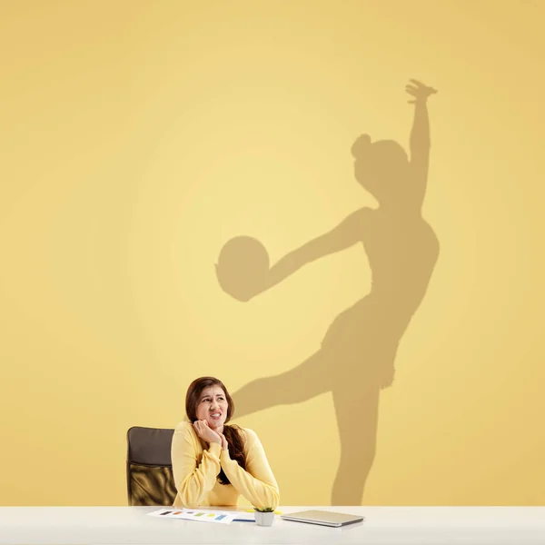 Giovane donna sognando il futuro nel grande sport durante il suo lavoro in ufficio. Diventare una leggenda. Ombra di sogni sul muro dietro di lei. Copyspace. — Foto Stock