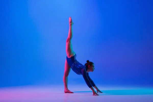 Young flexible girl isolated on blue studio background. Young female model practicing artistic gymnastics. Exercises for flexibility, balance.