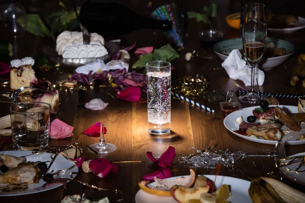 Early morning after the party. Glass of pure, cold water on the table with confetti and serpentine, leftovers, flower petals