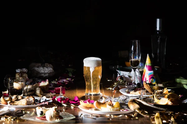 Early morning after the party. Glass of light, cold lager, beer on the table with confetti and serpentine, leftovers, flower petals